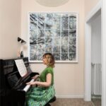 Žena sedí pri čiernom klavíri pod vysokým stropom so svetlou lampou nad terazzovou podlahou.