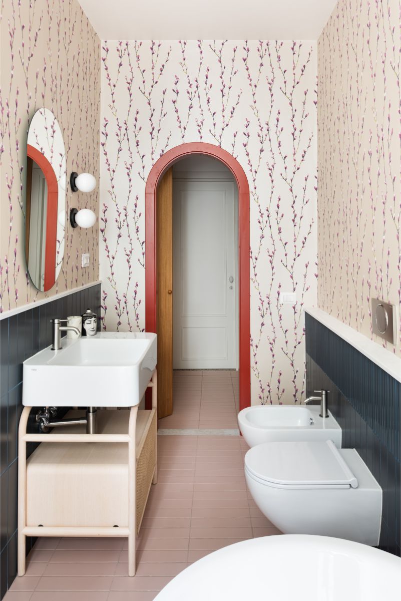 Kúpeľňa s ružovou dlažbou a tapetou so vzorom konárikov a tmavomodrými obkladmi. a oblúkový priechod s červeným lemom.
