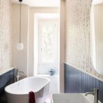 Biela voľne stojaca vaňa a obdĺžnikové umývadlo sú v kúpeľni s tapetou s rastlinným vzorom a tmavomodrými obkladmi.