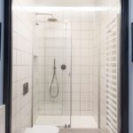 Sprchový kút s bielymi štvorcovými obkladmi a sklenenou stenou v kúpeľni so vzorovanou dlažbou a modrými stenami.