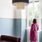 Žena v ružovom župane stojí pri okne v miestnosti s modrým farebným pásom na stene, terazzovou podlahou a fialovým ležadlom.