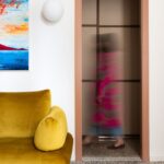 Horčicovo žltá zamatová sedačka pri stene s výrazným abstraktným obrazom.