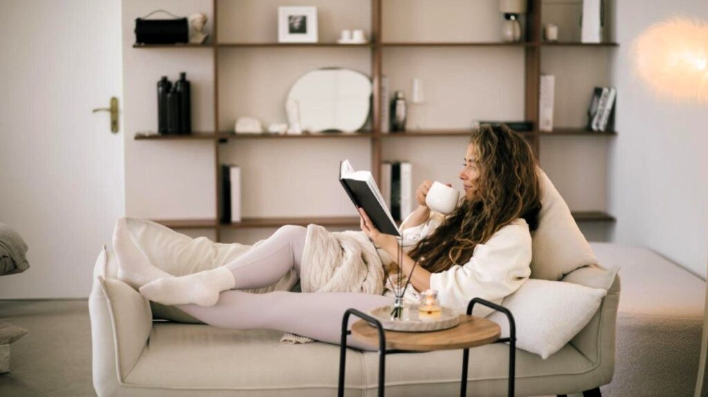 Žena leží na sedačke s hrnčekom v ruke a číta knihu.