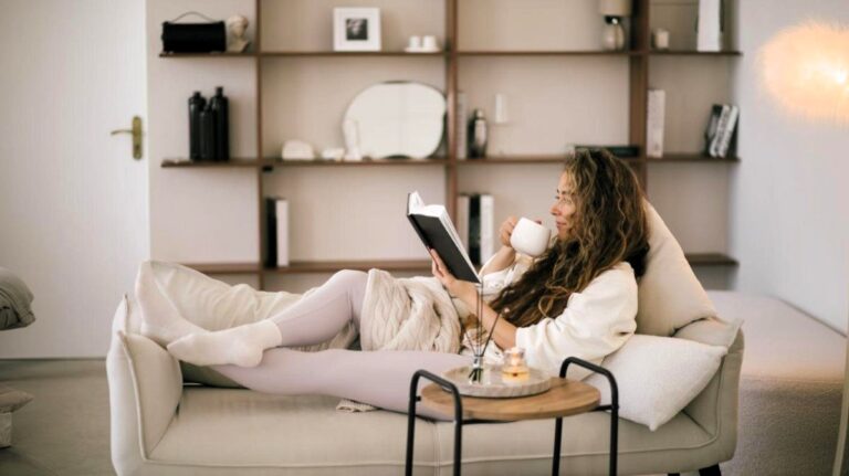 Žena leží na sedačke s hrnčekom v ruke a číta knihu.