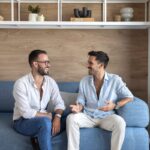 Dvaja muži sedia na modrej sedačke pred stenou obloženou svetlým drevom.