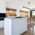 Moderná kuchyňa s bielym ostrovčekom, vstavanými spotrebičmi a dreveným obkladom, doplnená vysokými drevenými stoličkami.