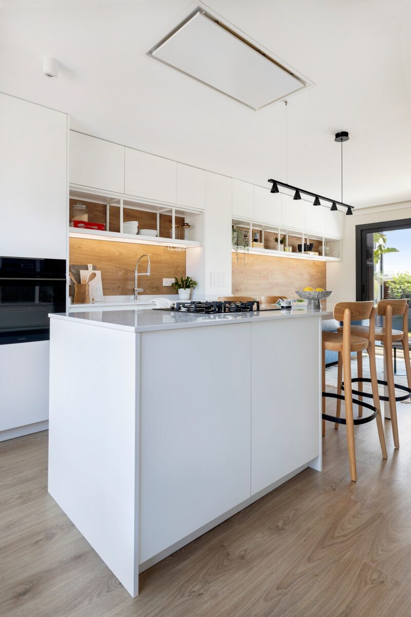 Moderná kuchyňa s bielym ostrovčekom, vstavanými spotrebičmi a dreveným obkladom, doplnená vysokými drevenými stoličkami.