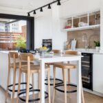 Biela kuchyňa s varným ostrovčekom, vysokým jedálenským stolom a drevenými barovými stoličkami.