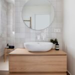 Kúpeľňa s bielymi štvorcovými obkladačkami, drevenou skrinkou a oválnym umývadlom pod podsvieteným zrkadlom.