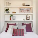 Spálňa s bielou posteľou, vankúšmi v červenej a fialovej farbe a bielym vstavaným policovým systémom.