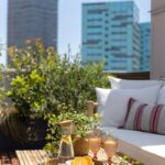 Bambusový stolík s nápojom a ovocím na terase s rastlinami a bielou pohovkou, s výhľadom na moderné budovy.