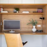 Moderný pracovný kútik so vstavaným stolom z dreva, bielymi skrinkami a dizajnovou stoličkou.