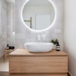 Kúpeľňa s bielymi štvorcovými obkladačkami, drevenou skrinkou a oválnym umývadlom pod podsvieteným zrkadlom.