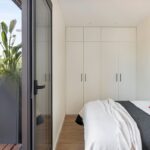 Svetlá minimalistická spálňa s bielou vstavanou skriňou, drevenou podlahou a posuvnými dverami na terasu.
