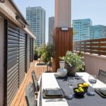 Terasa s drevenou podlahou, jedálenským setom s tmavosivými stoličkami a výhľadom na moderné mrakodrapy.