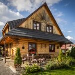 Drevený zrubový dom s čiernou strechou a vonkajším posedením obklopený záhradou a zeleňou.