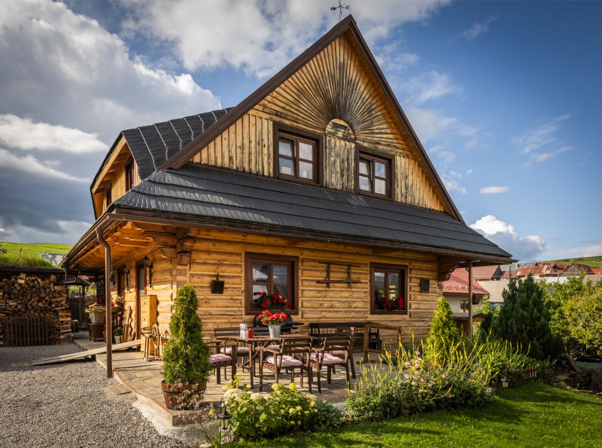 Drevený zrubový dom s čiernou strechou a vonkajším posedením obklopený záhradou a zeleňou.