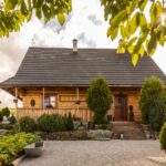 Drevený zrubový dom s tmavou šindľovou strechou a upravenou predzáhradkou s kríkmi a štrkovou plochou.