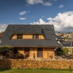 Drevená zrubová chalupa s čiernou strechou a kopou naskladaného dreva pred dedinskou zástavbou.