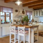 Biela kuchynská linka s drevenou doskou, ostrovčekom s dvoma barovými stoličkami a trámovým dreveným stropom.