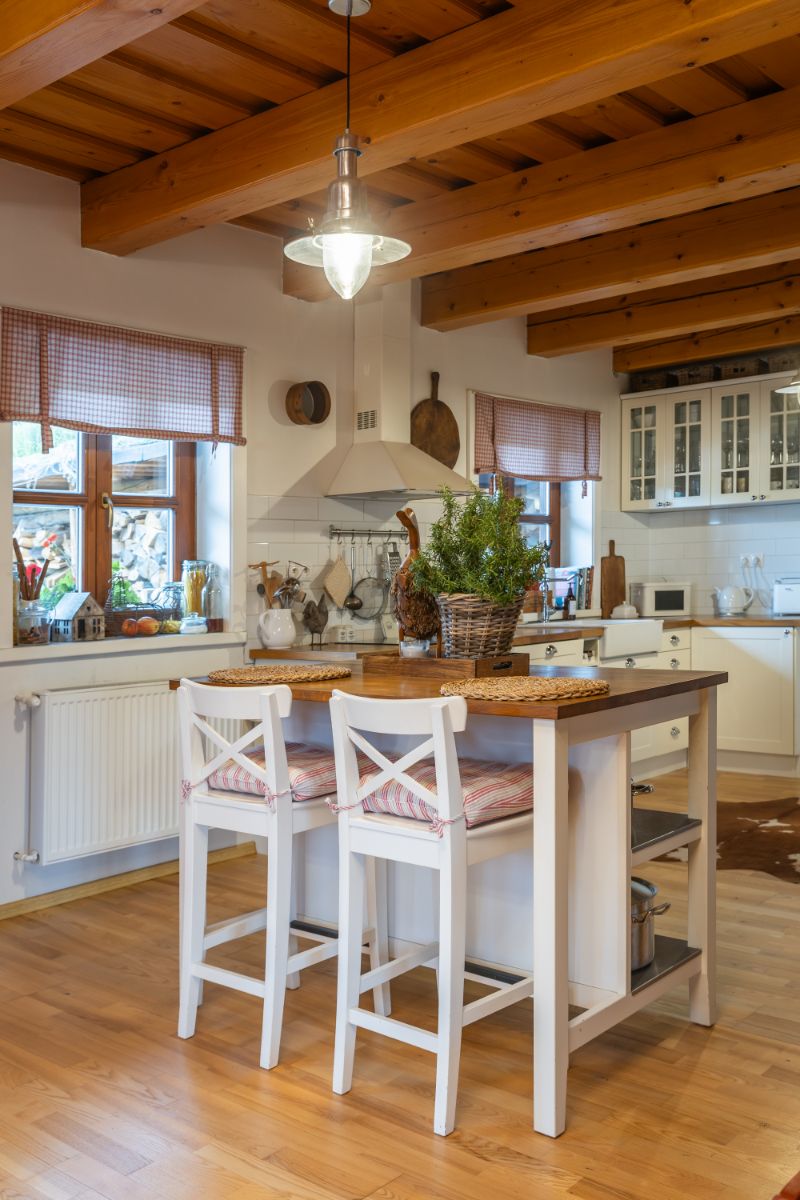 Biela kuchynská linka s drevenou doskou, ostrovčekom s dvoma barovými stoličkami a trámovým dreveným stropom.