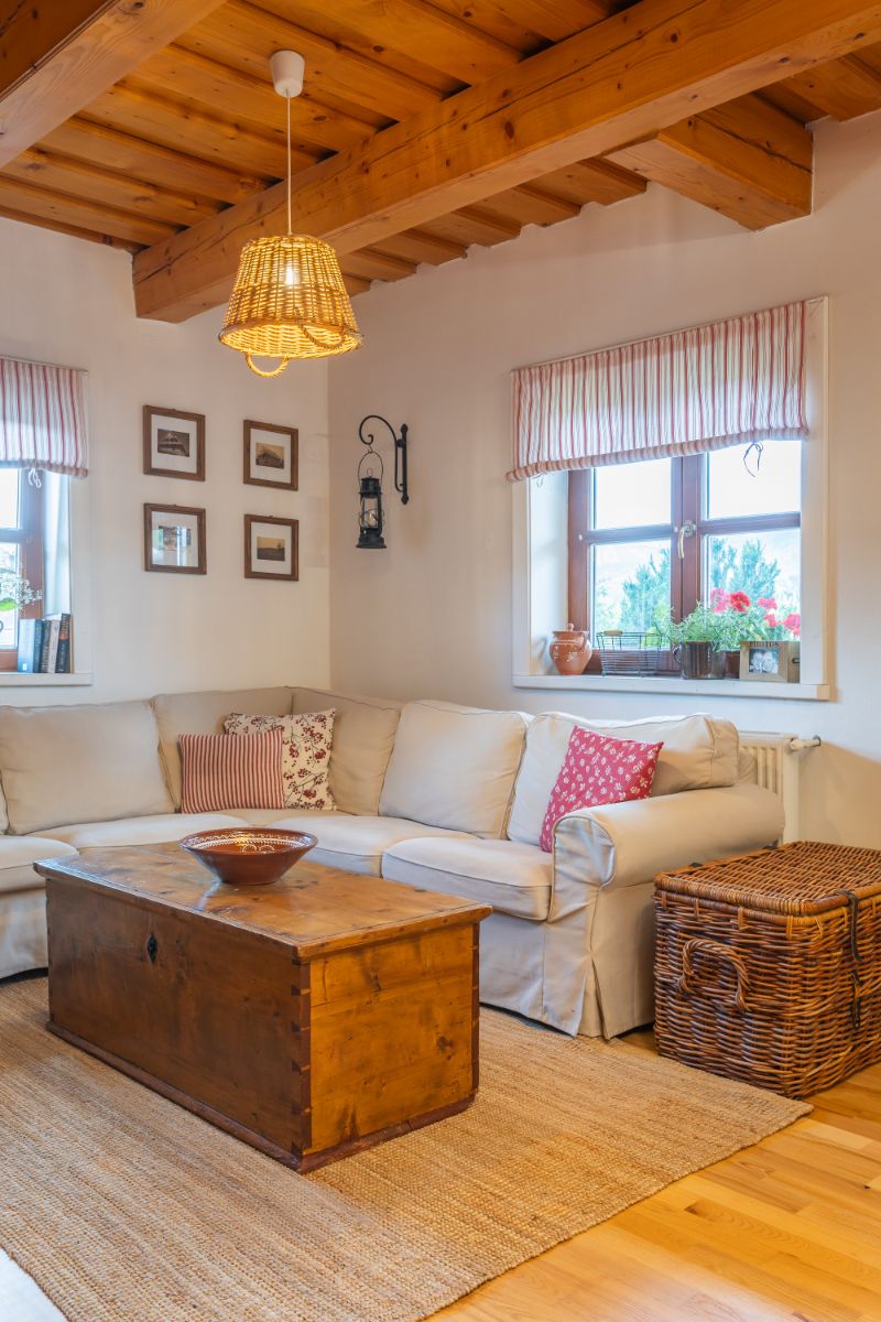 Rohová biela sedacia súprava, drevená truhlica ako konferenčný stolík a prútený kôš v obývačke s trámovým stropom.