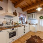 Vidiecka biela kuchyňa s drevenou pracovnou doskou, indukčnou varnou doskou a digestorom pod drevenými trámami.