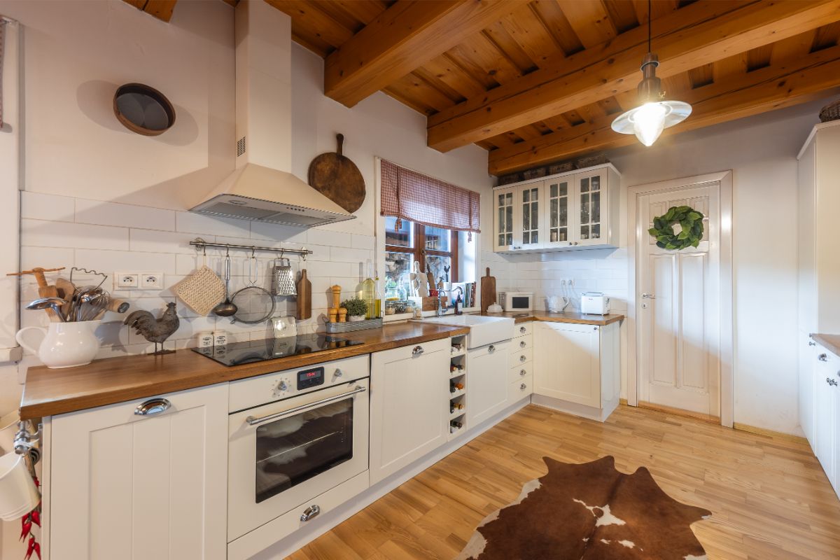 Vidiecka biela kuchyňa s drevenou pracovnou doskou, indukčnou varnou doskou a digestorom pod drevenými trámami.