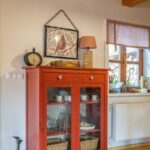 Červená presklená komoda s bielym riadom, starožitnými hodinami a obrazom vtáka v rámčeku.