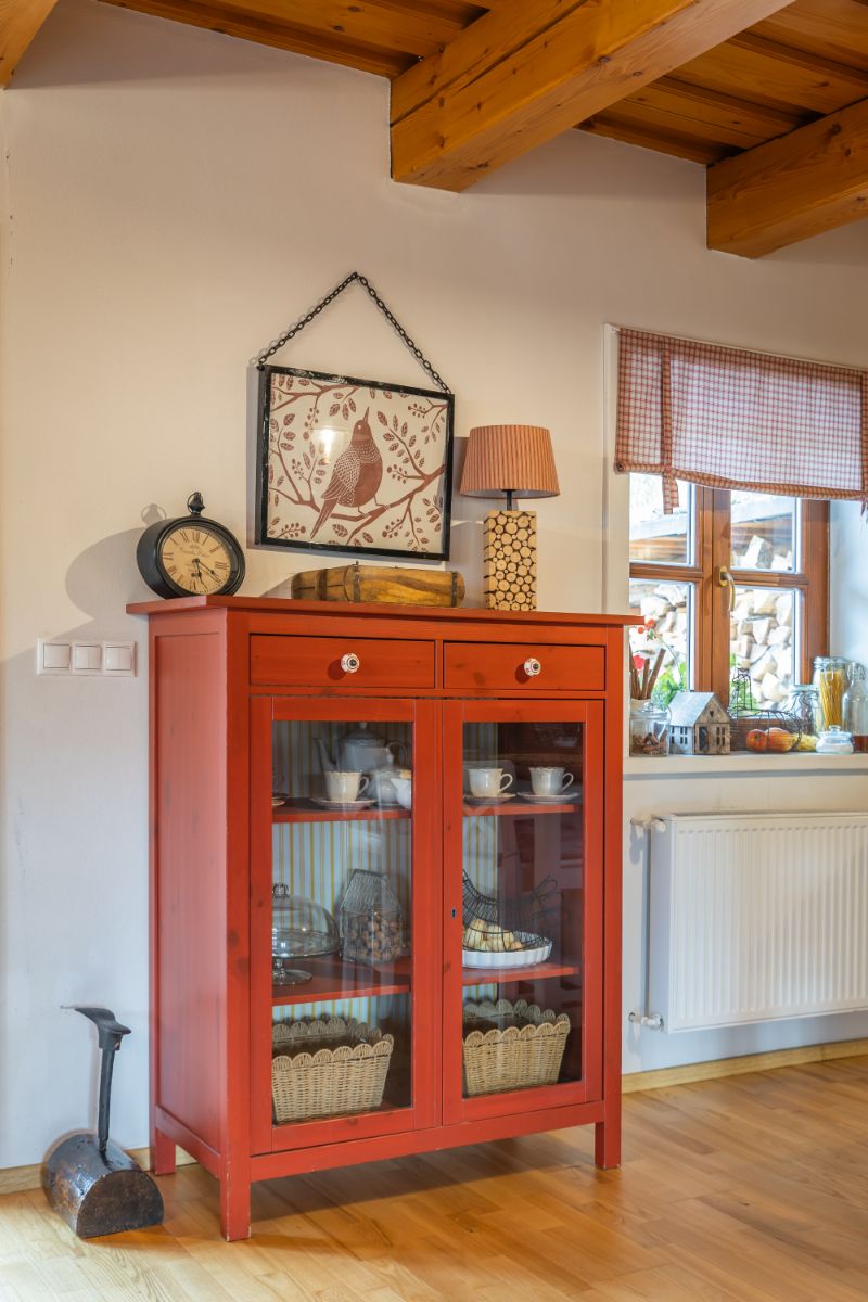 Červená presklená komoda s bielym riadom, starožitnými hodinami a obrazom vtáka v rámčeku.