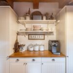 Biela kuchynská skrinka s drevenou doskou, nápisom Bakery a otvorenými policami s rustikálnym príslušenstvom.