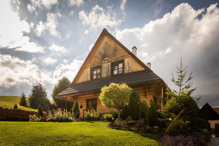 Drevený zrubový dom s čiernou strechou a vonkajším posedením obklopený záhradou a zeleňou.