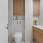 Minimalistická kúpeľňa s bielym WC, dreveným lamelovým obkladom a svetlosivými betónovými stenami.