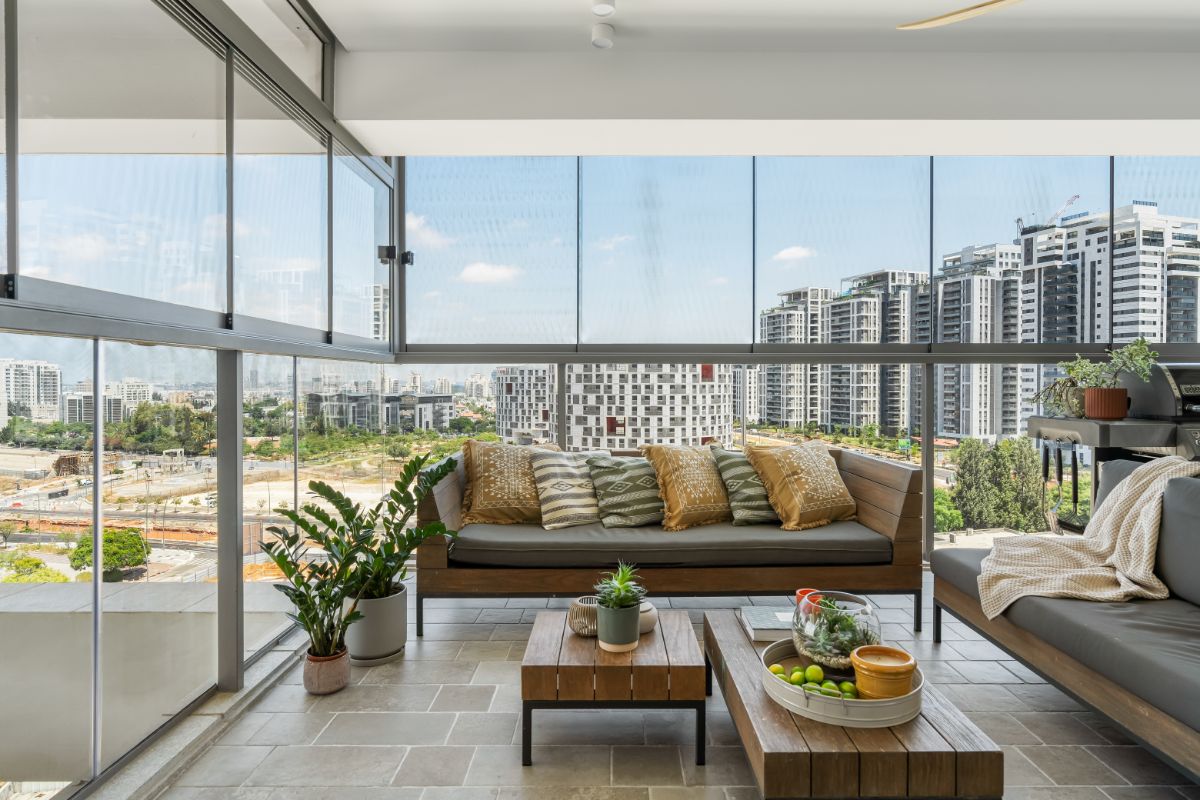 Presklená terasa s tmavým dreveným záhradným nábytkom, vankúšmi a výhľadom na mestské budovy.