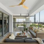 Terasová obývačka s drevenou sedacou súpravou, šedými vankúšmi a dreveným stropným ventilátorom.