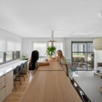 Otvorená kuchyňa a obývačka s dlhým dreveným ostrovčekom, bielymi skrinkami a veľkými lampami.