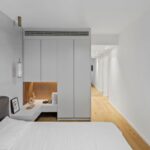 Spálňa s bielou posteľou, svetlosivými skriňami a nočným stolíkom s dreveným podsvieteným výklenkom