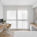 Svetlá domáca kancelária a spálňa s dlhým dreveným stolom, ergonomickou stoličkou a bielymi priesvitnými závesmi.