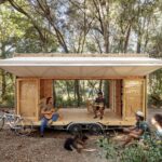 Drevená terasa a interiér mobilného domu ako pódium pre dvoch hudobníkov, s bicyklom a poslucháčmi na lesnej pôde.