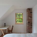 Svetlá podkrovná spálňa s bielou zošikmenou stenou, malým oknom, tehlovým komínom a minimalistickým stolíkom.