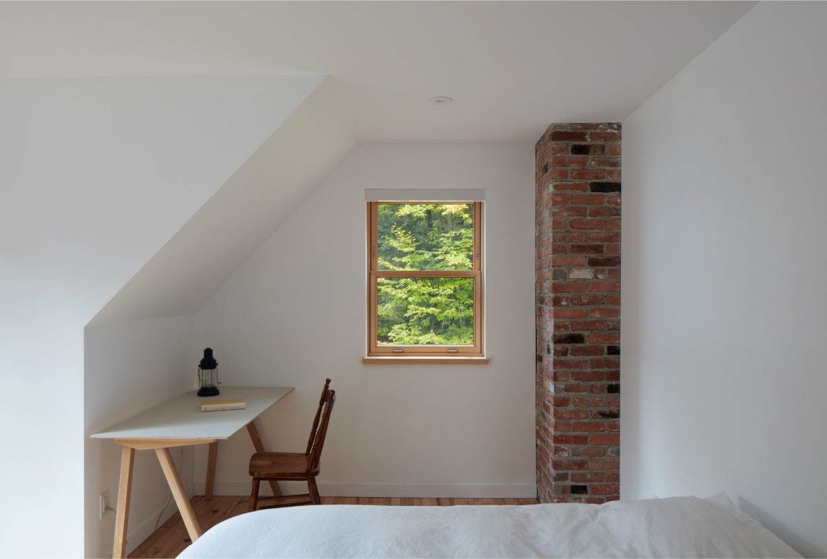 Svetlá podkrovná spálňa s bielou zošikmenou stenou, malým oknom, tehlovým komínom a minimalistickým stolíkom.