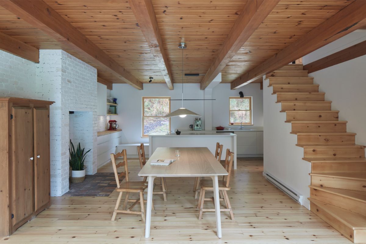 Jedáleň a kuchyňa s dreveným trámovým stropom, bielou kuchynskou linkou, drevenou skriňou a schodmi na poschodie.