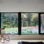 Veľkoformátpvé okná s čiernym rámom s výhľadom na les, hojdacie kreslo v rohu.