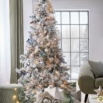 Biely zasnežený vianočný stromček s jemnými modro-bielymi ozdobami a teplým osvetlením pri okne v interiéri.