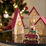 Drevené svietiace domčeky s červenými strechami a drevené auto s vianočným stromčekom na lesklom stole.