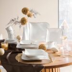 Okrúhly drevený stôl so slávnostným prestieraním z bieleho porcelánu so zlatým okrajom a zlatými príbormi.