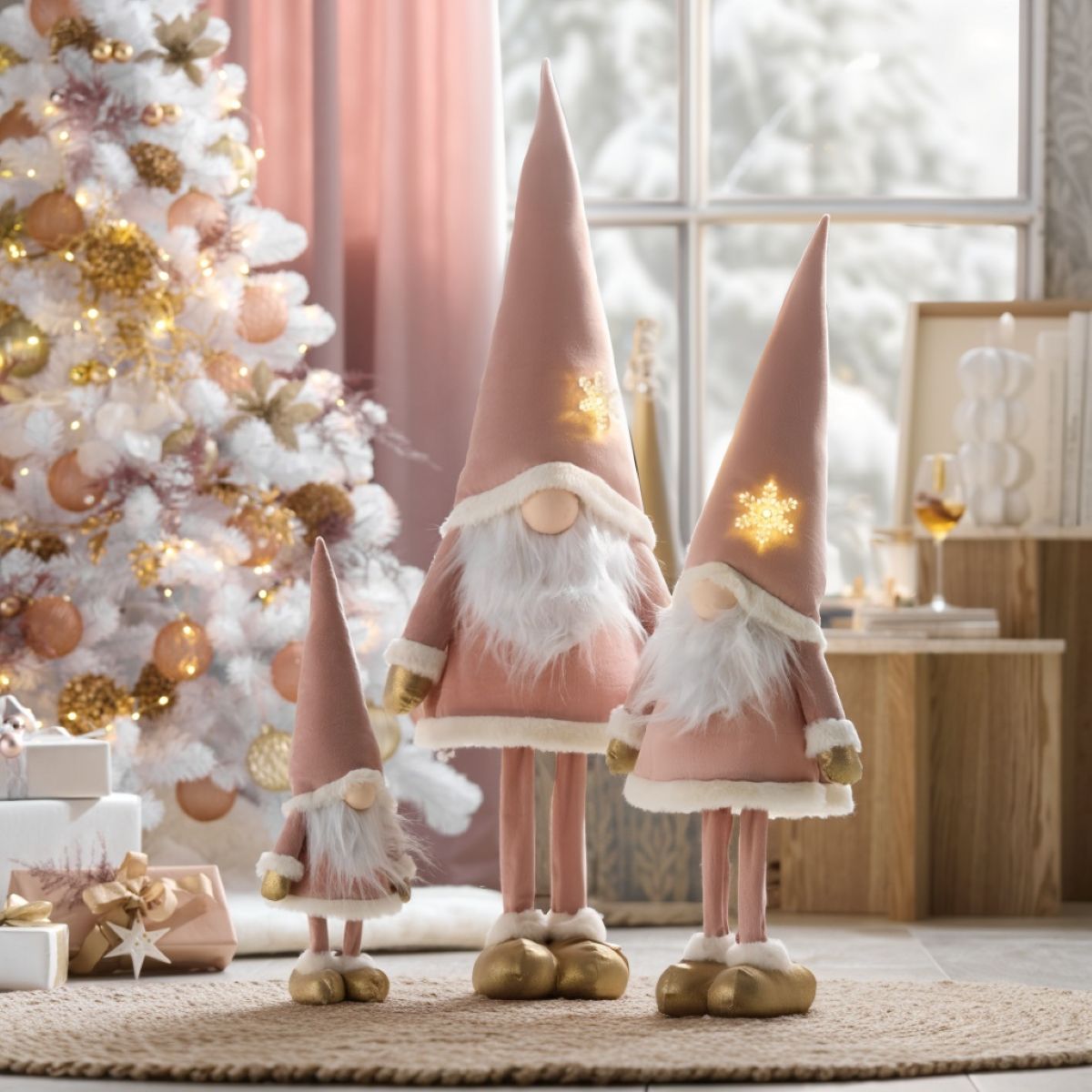 rojica ružovo-zlatých látkových vianočných škriatkov s bielymi bradami a svietiacimi hviezdami pri bielom stromčeku.