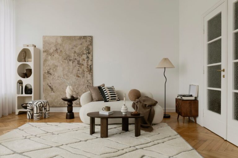 Obývačka v neutrálnych tónoch s bielou bouclé sedačkou, abstraktným obrazom a okrúhlym dreveným konferenčným stolíkom.