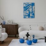 Moderná obývačka s bielym bouclé nábytkom, tmavou komodou a skleneným stolíkom s kobaltovo modrými nohami.