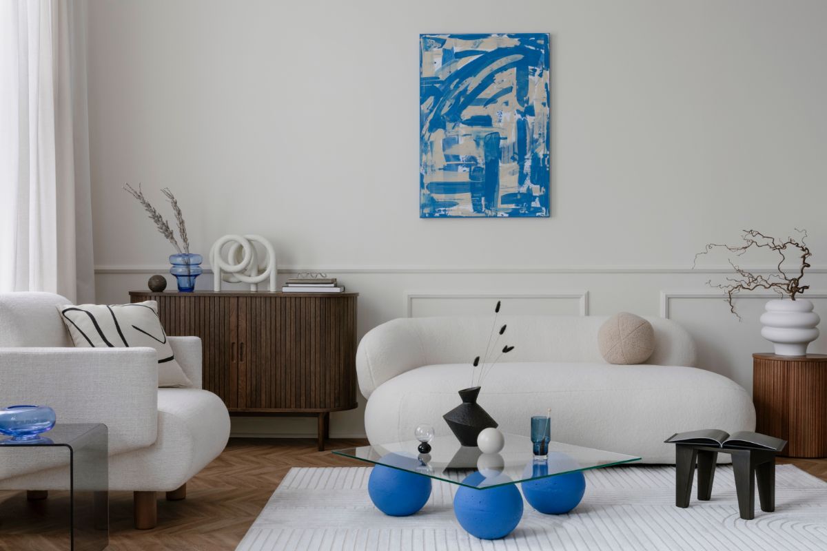 Moderná obývačka s bielym bouclé nábytkom, tmavou komodou a skleneným stolíkom s kobaltovo modrými nohami.